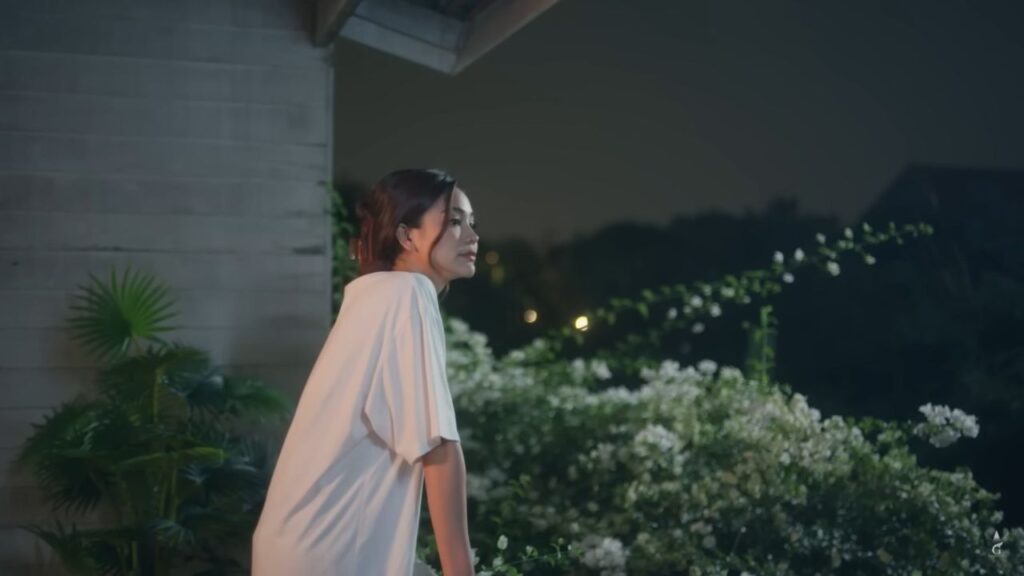 A sapphic character from Show Me Love, a Thai GL drama, stands alone on her terrace, her face half-lit by soft evening light, reflecting inner conflict and identity.